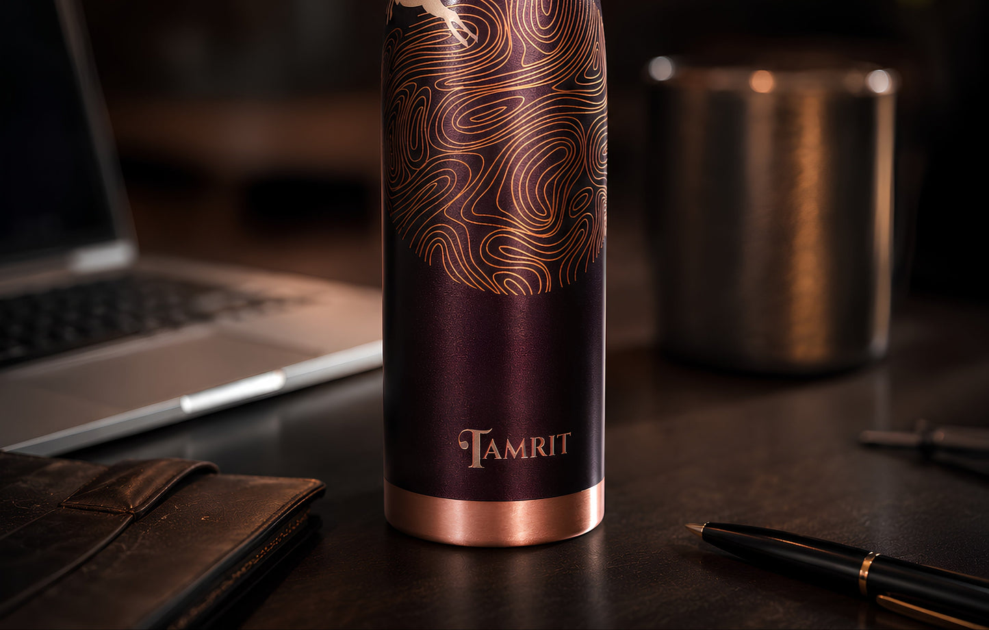 A dark plum purple insulated water bottle with a rose gold cap and base, featuring a wave pattern and a deer graphic, standing on a dark desk with a laptop and other office items blurred in the background.